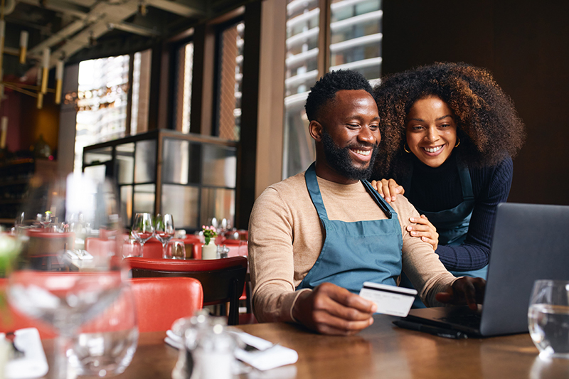 Couple smiling looking at credit card and computer sitting at a table inside of an empty restaurant