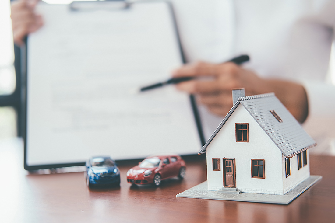 Dark oak red table with a toy replica house and toy replica cars, a blurred picture of a man in a white dress shirt in the background holding up a document and pen.