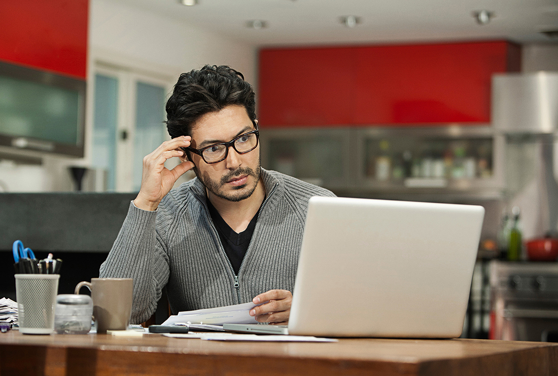 Middle aged man sitting at table in his kitchen with his laptop open looking frustrated