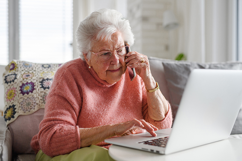 Elderly lady sitting on couch talking on phone while looking at her laptop