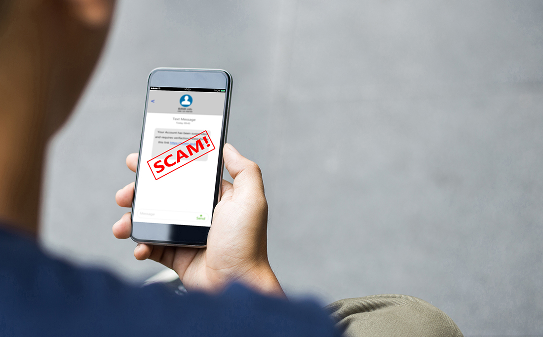 A man in a navy blue shirt looks down at his cell phone, which shows he received a text message that includes a blue hyperlink. The word ‘scam’ is written in bold, red, capital letters over the text message.
